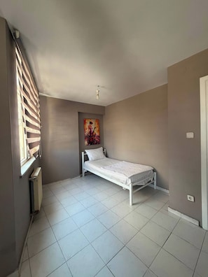 A small, single-bed room with a radiator, a window with blinds, and a painting on the wall.