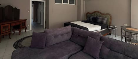 Ein Wohnzimmer mit einem grauen Ecksofa, einem kleinen runden Couchtisch und einem Bett mit Kopfteil.