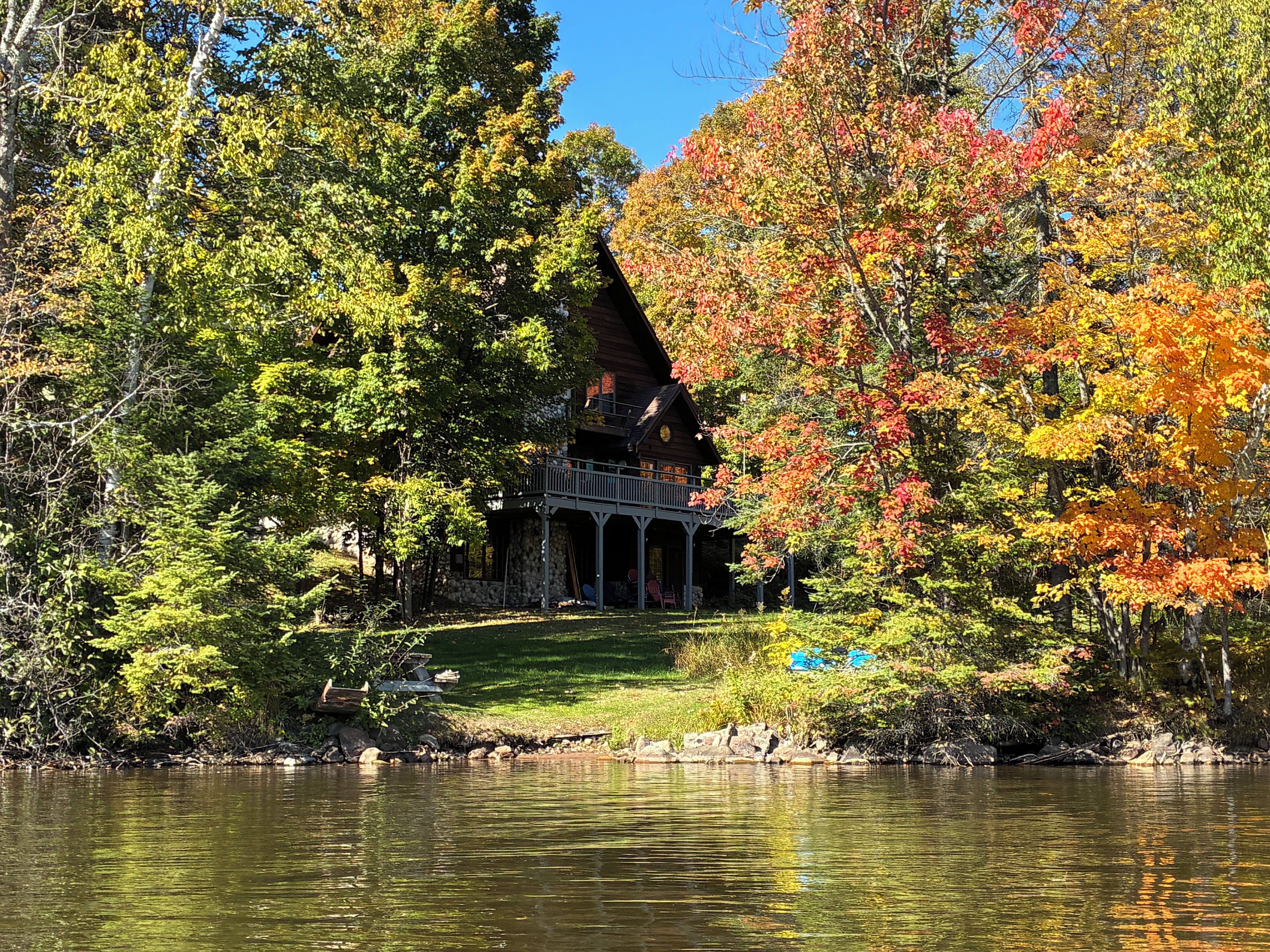 A lakeside cabin with a porch, surrounded by autumn foliage and a calm lake.