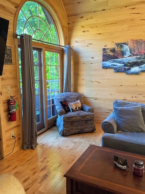 Interior acogedor de una cabaña con piso de madera, un sofá, un sillón con almohadones decorativos, una mesa de centro y un gran ventanal con cortinas.