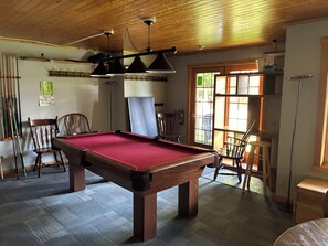 Ein Billardtisch in einem Raum mit Holzboden und -wänden, ein Sessel und eine Tür, die nach draußen führt.
