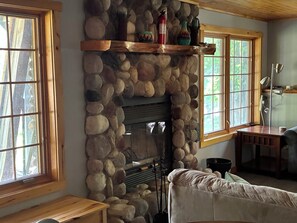 Una chimenea de piedra con repisa de madera, un sofá y una ventana con vista a árboles.