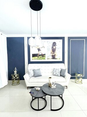 Un salon avec un canapé blanc, deux tables rondes, un luminaire suspendu et un tableau encadré au mur.