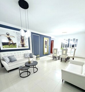 A modern living room with a white sofa, a round coffee table, and a dining area with chairs and a table. - Spacious 3BR Near Airport & Ocean (Santo Domingo Este)