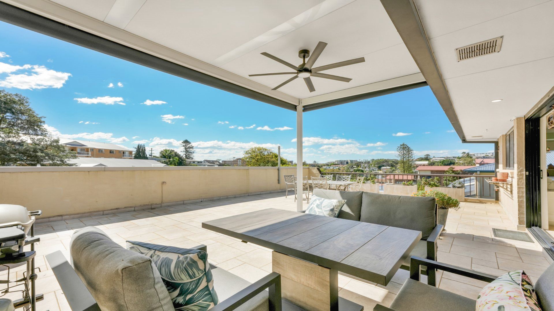 A covered outdoor patio with a table and chairs, a ceiling fan, and a view of the surrounding neighborhood.