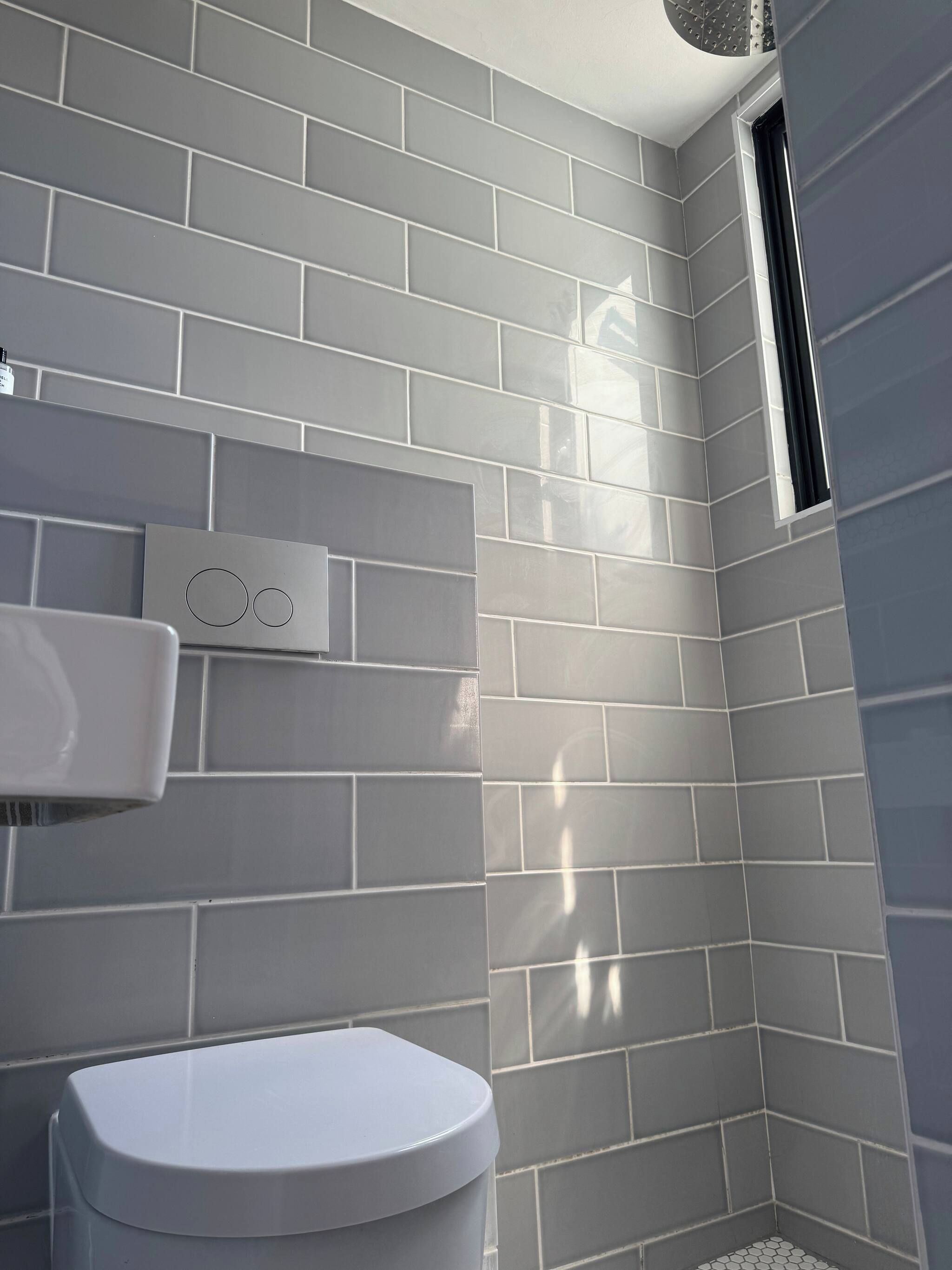 A bathroom with grey tiled walls, a white toilet, and a window.
