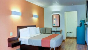 Quarto de hotel com cama, mesa de cabeceira, pequena mesa com uma cadeira, espelho e área de cozinha compacta.