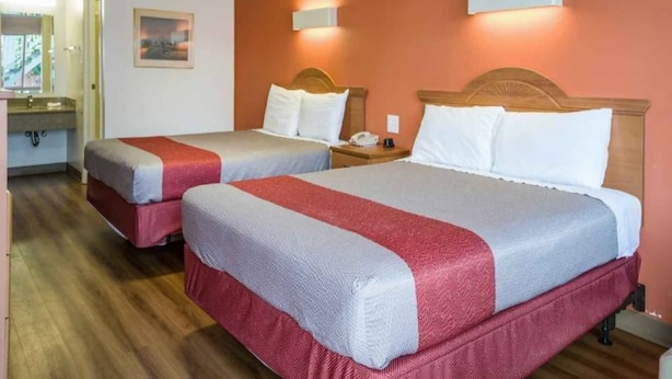 Quarto de hotel com duas camas, cabeceira de madeira, mesa de cabeceira e luminária fixada na parede.