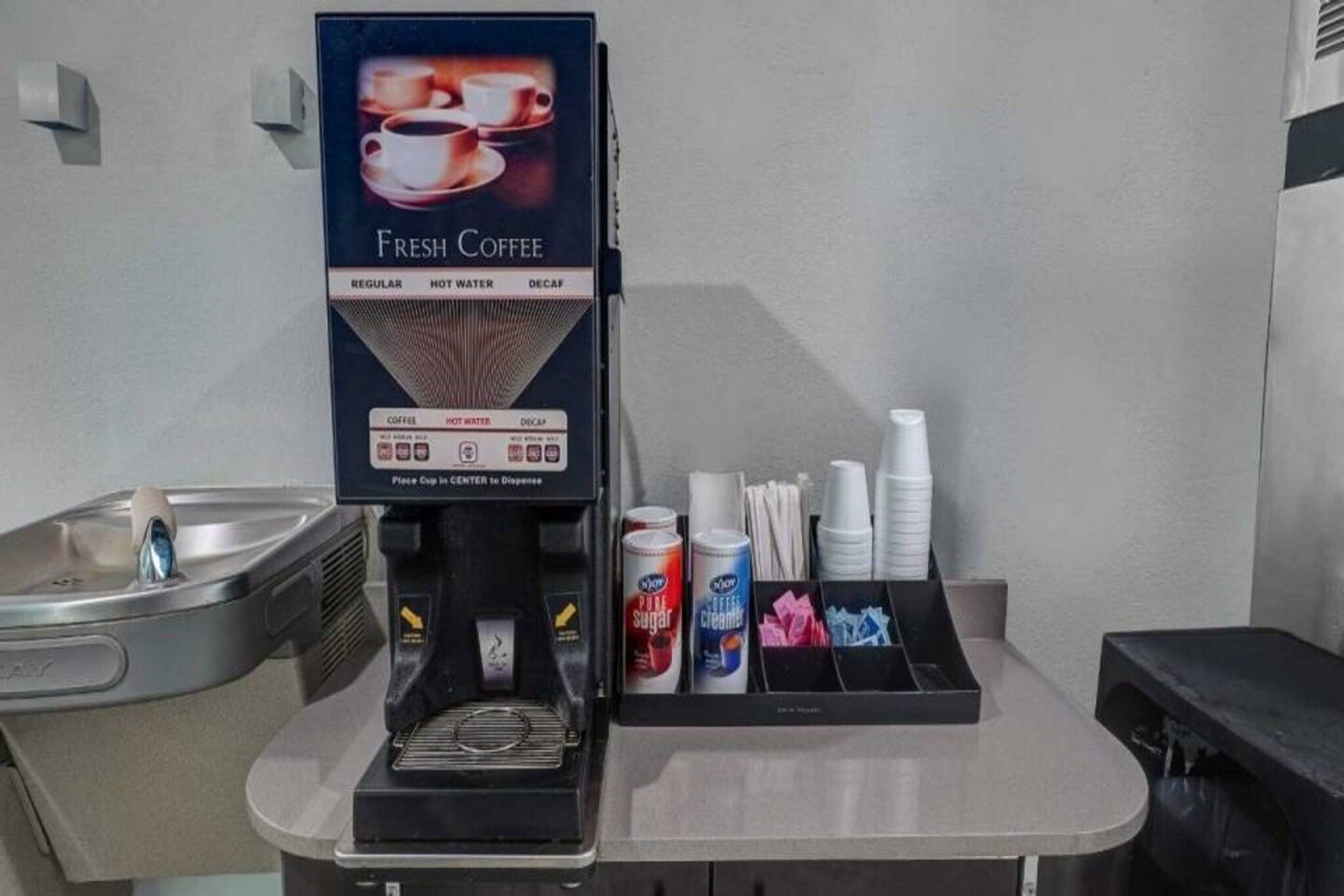Eine Kaffeemaschine mit einem Schild, das ‚Frischer Kaffee‘ anzeigt, ein Becherautomat und eine Kaffeestation mit Bechern, Deckeln und Sahneportionen.