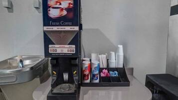 Eine Kaffeemaschine mit einem Schild, das ‚Frischer Kaffee‘ anzeigt, ein Becherautomat und eine Kaffeestation mit Bechern, Deckeln und Sahneportionen.