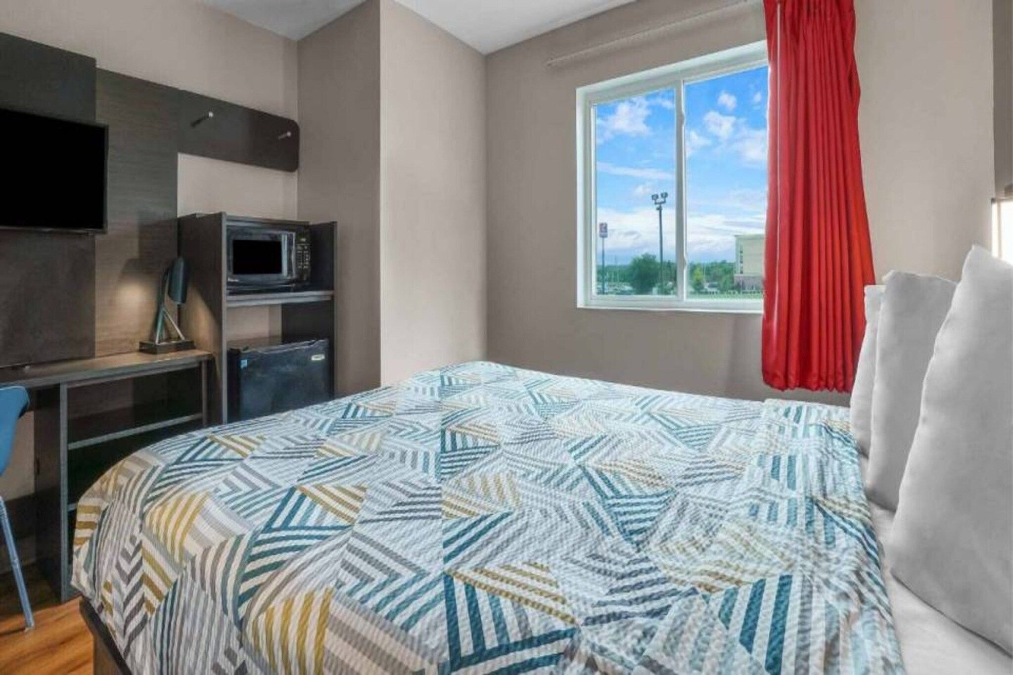 A bedroom with a bed, a desk with a computer and a microwave, a window with a view of the sky, and a red curtain.