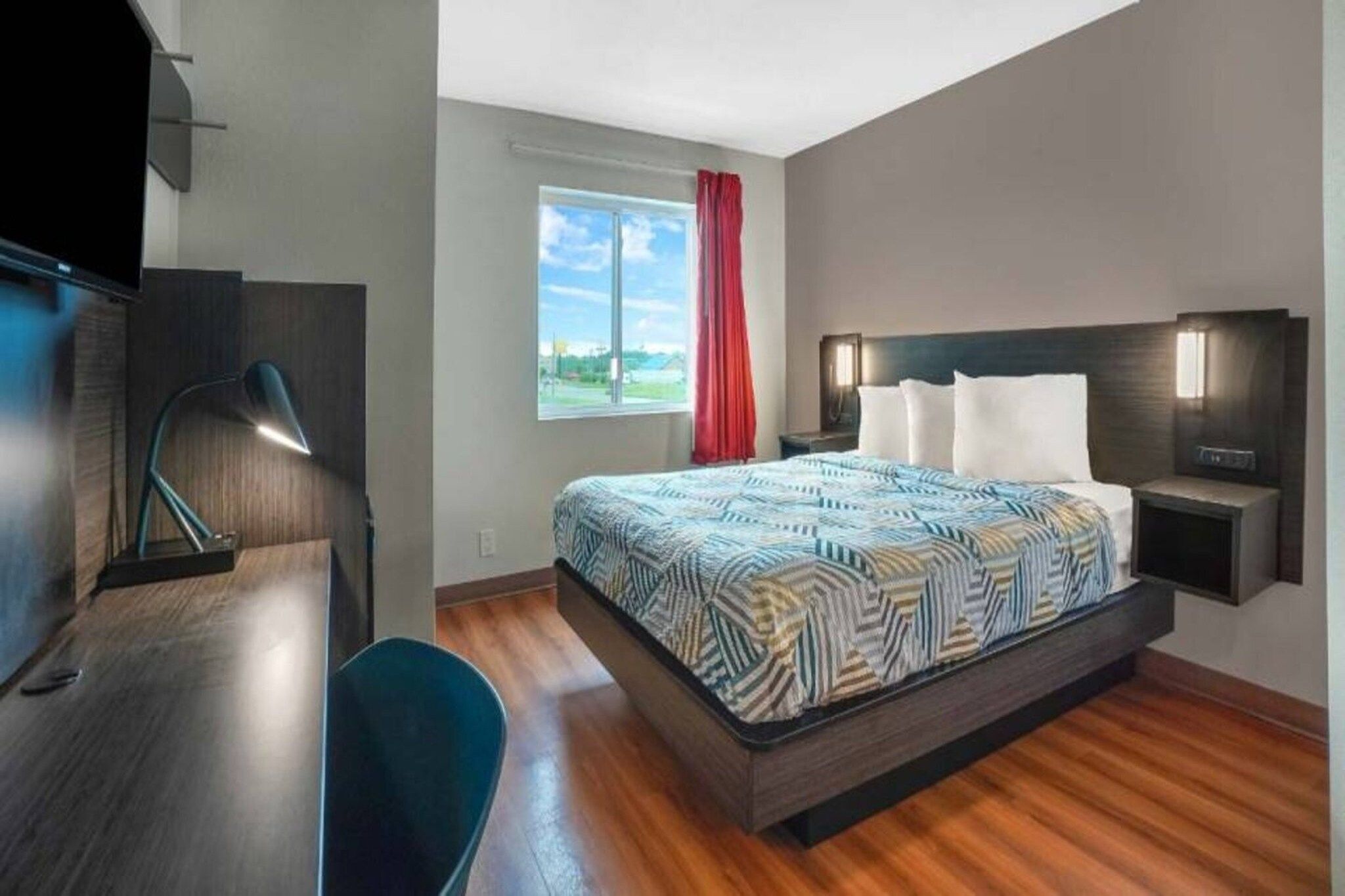 A hotel room with a bed, bedside tables, a desk, a chair, a window with a view, and a red curtain.