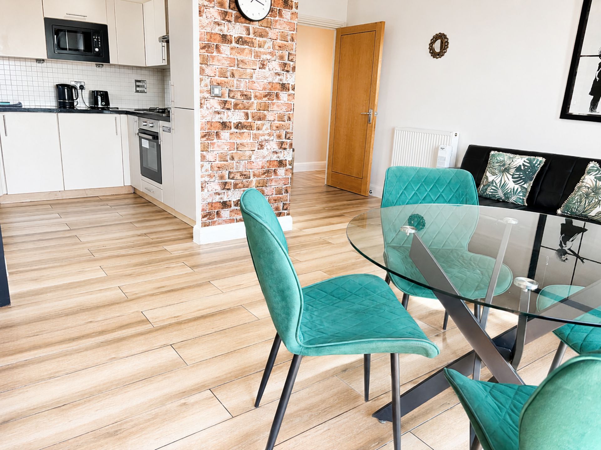 Una cocina moderna con armarios blancos, pared de acento de ladrillo y comedor con sillas color turquesa y mesa de vidrio.