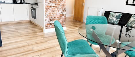Una cocina moderna con armarios blancos, pared de acento de ladrillo y comedor con sillas color turquesa y mesa de vidrio.