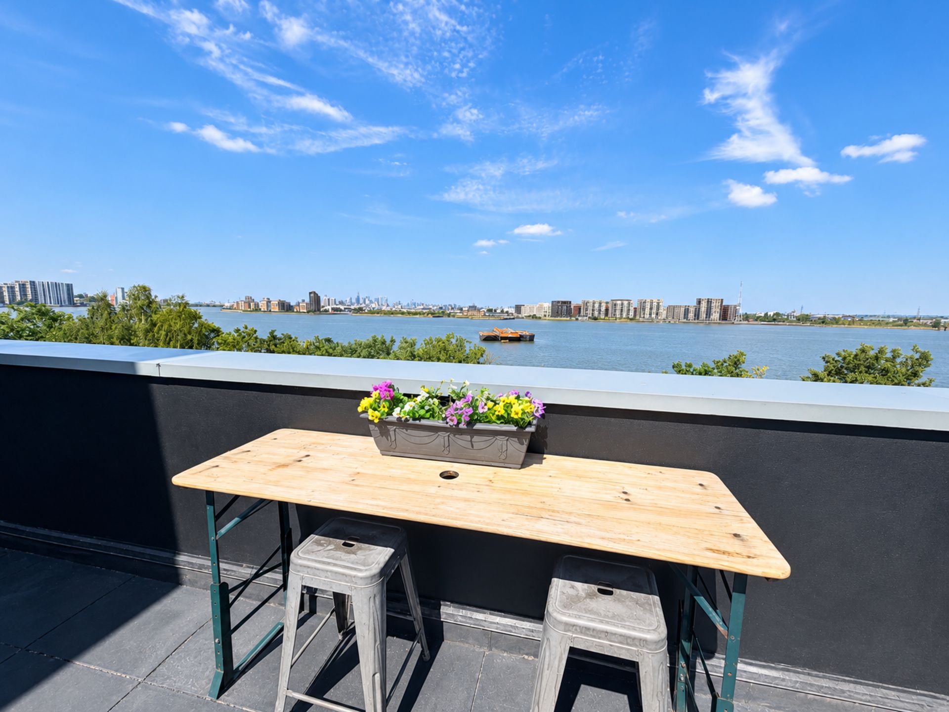 A wooden table with a flower planter on a balcony overlooking a river with buildings in the distance.