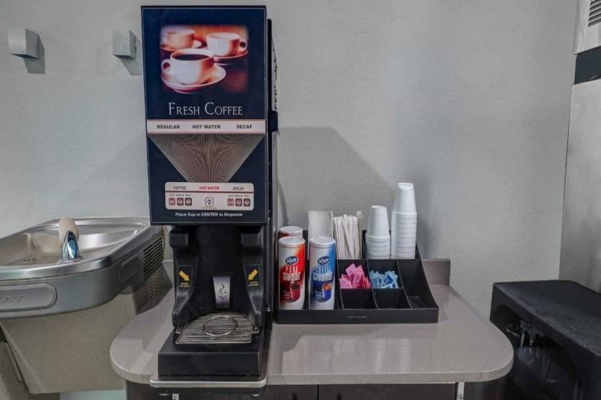 A coffee machine with a fresh coffee sign, a water dispenser, and a cup holder with disposable cups.