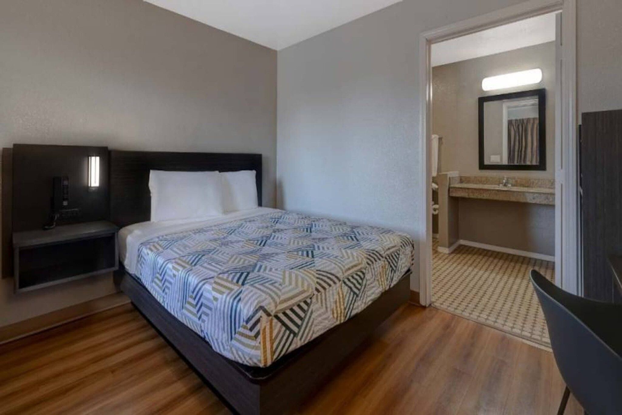 Habitación de hotel con cama, mesita de noche, una silla, un escritorio pequeño y un baño con espejo y lavamanos.