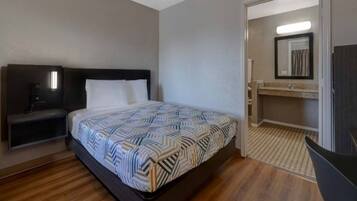 Habitación de hotel con cama, mesita de noche, una silla, un escritorio pequeño y un baño con espejo y lavamanos.
