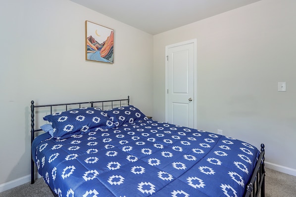 A single bed with a patterned blue bedspread and pillows, a metal headboard, a white door, and a framed picture on the wall.