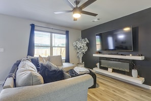 A modern living room with a flat-screen TV, a ceiling fan, and a large window with blinds.