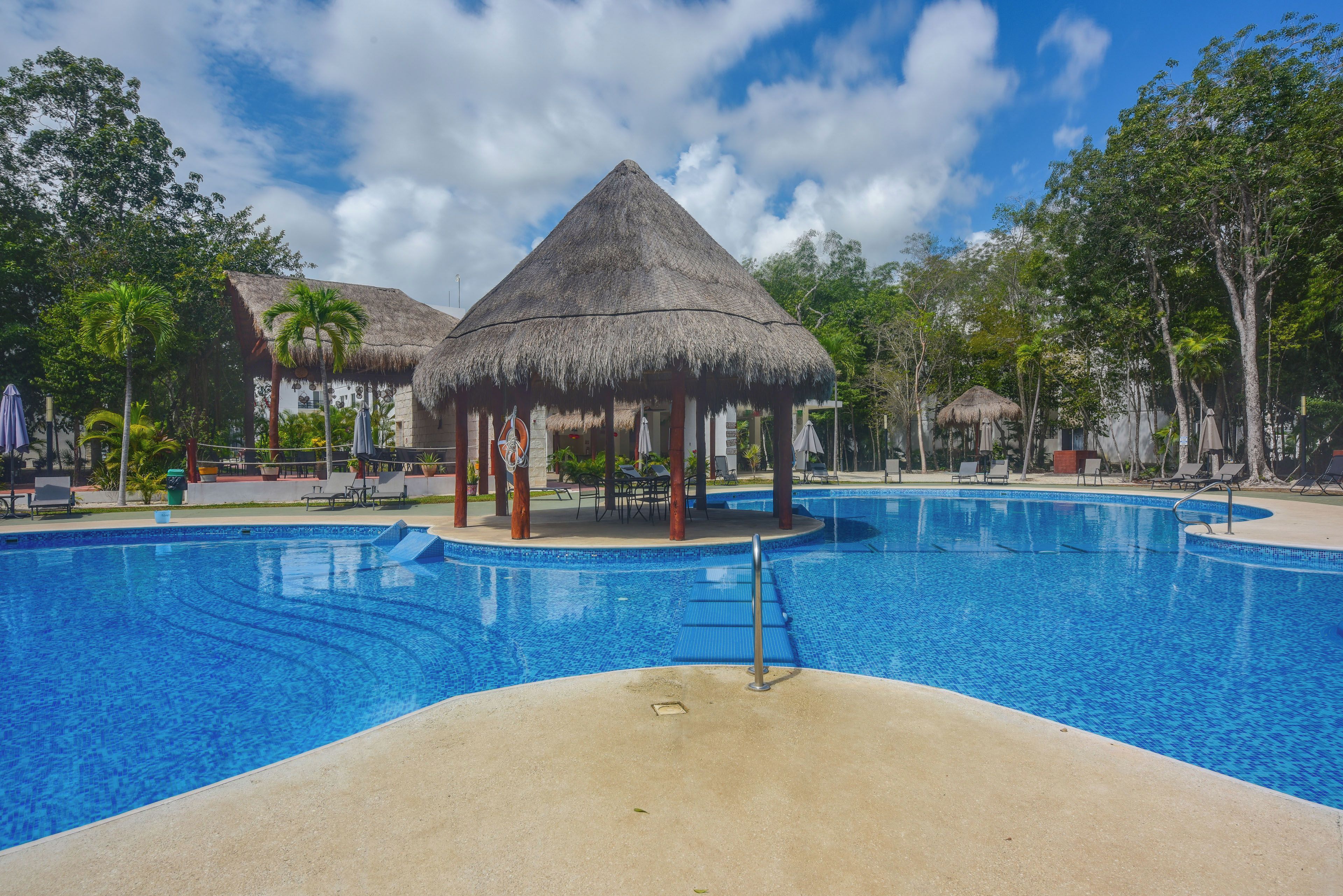 A swimming pool with a thatched-roof pavilion, lounge chairs, and palm trees.