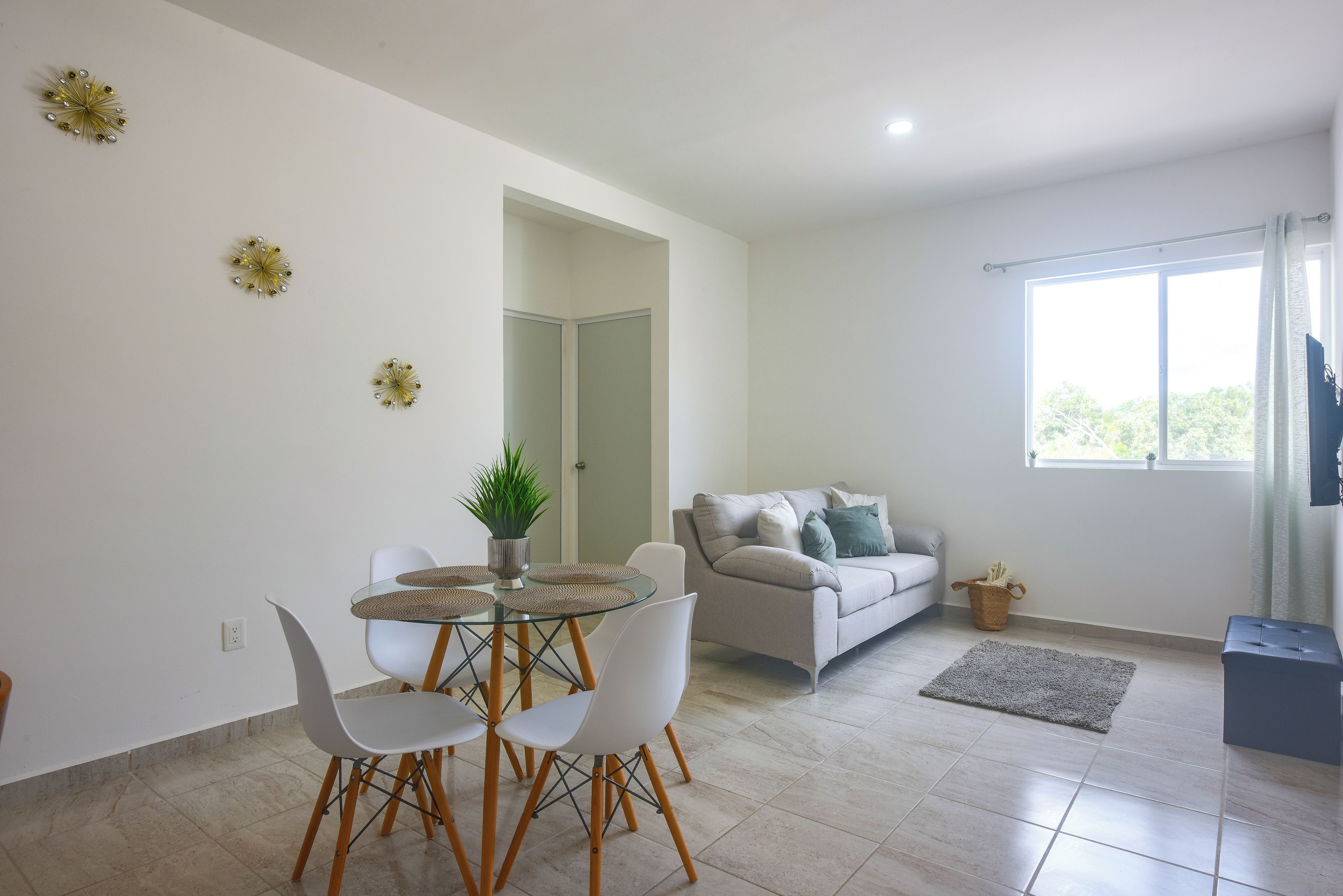 A modern living room with a glass dining table, white chairs, a grey sofa, and a large window with curtains.