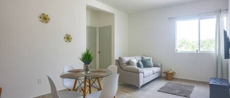 A modern living room with a glass dining table, white chairs, a grey sofa, and a large window with curtains.