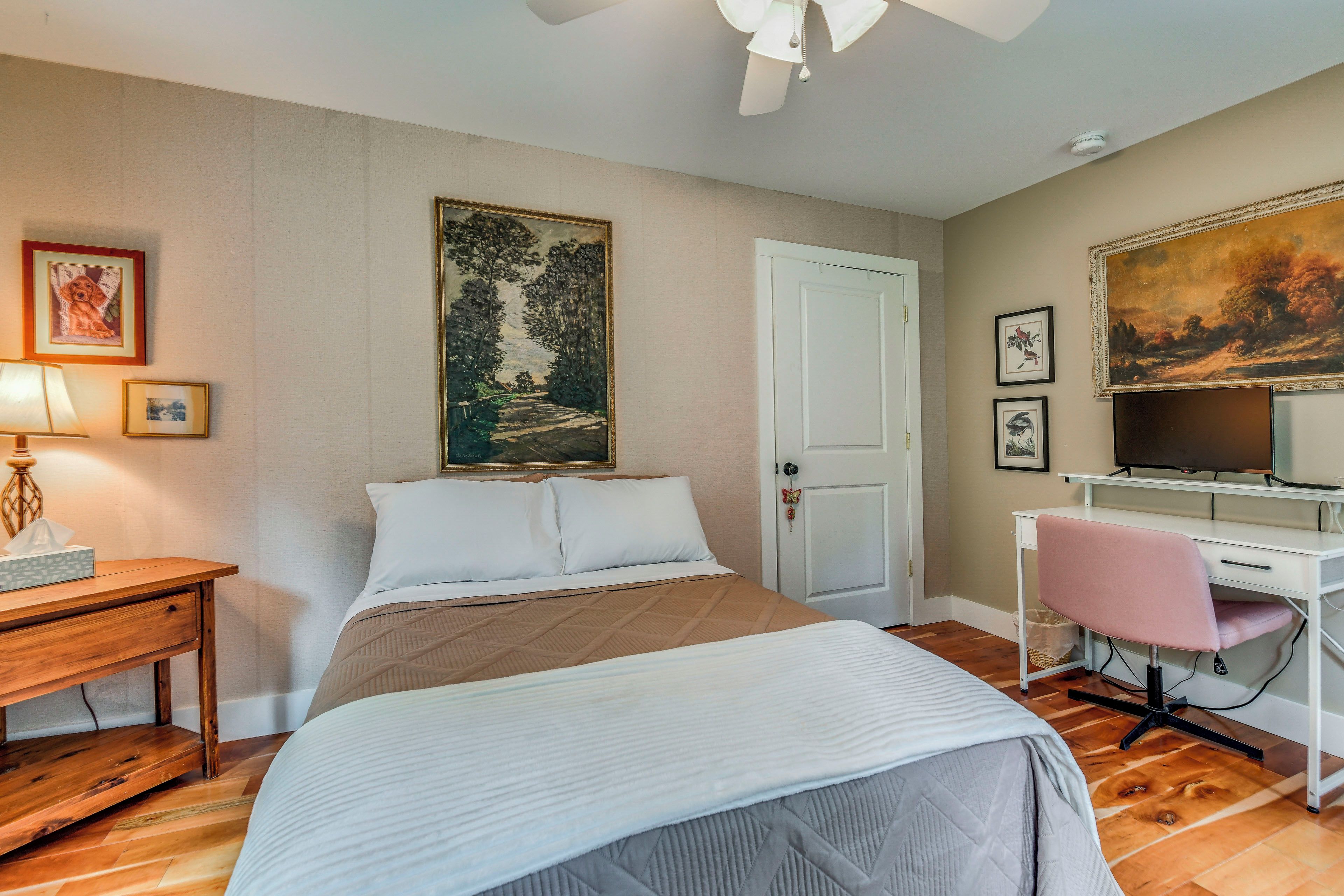 A bedroom with a bed, bedside table, lamp, desk with chair, and a painting on the wall.