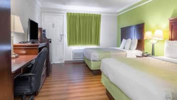 A hotel room with a desk, two beds, a window with green curtains, and a bed lamp.