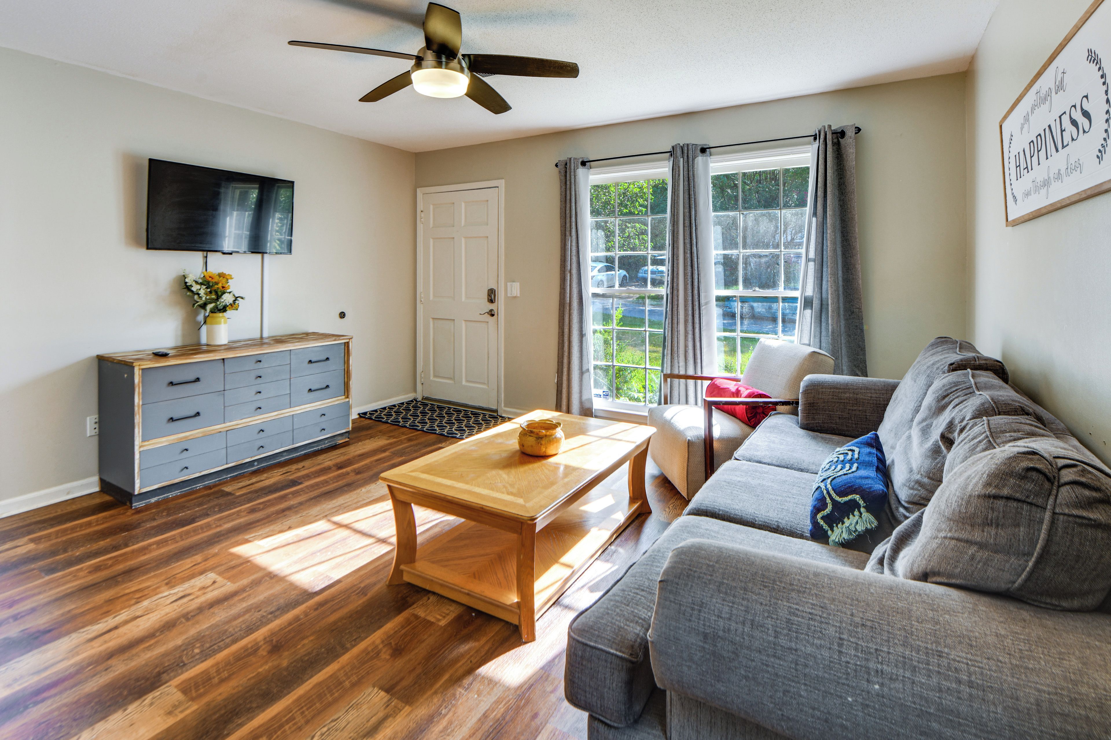 A living room with a wooden floor, a grey sofa, a wooden coffee table, a TV mounted on the wall, and a ceiling fan.