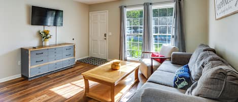A living room with a wooden floor, a grey sofa, a wooden coffee table, a TV mounted on the wall, and a ceiling fan.