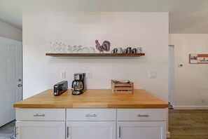 Una cocina con encimera de madera, cafetera, tostadora y estante con cristalería y un gallo decorativo.