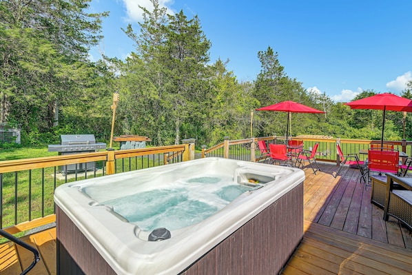 A hot tub on a wooden deck with a slide, surrounded by red chairs and umbrellas, and a barbecue grill in the background.