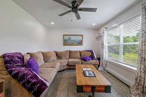 Una sala de estar con un sofá modular, una mesa de centro de madera, un ventilador de techo, un cuadro en la pared y una ventana con persianas.