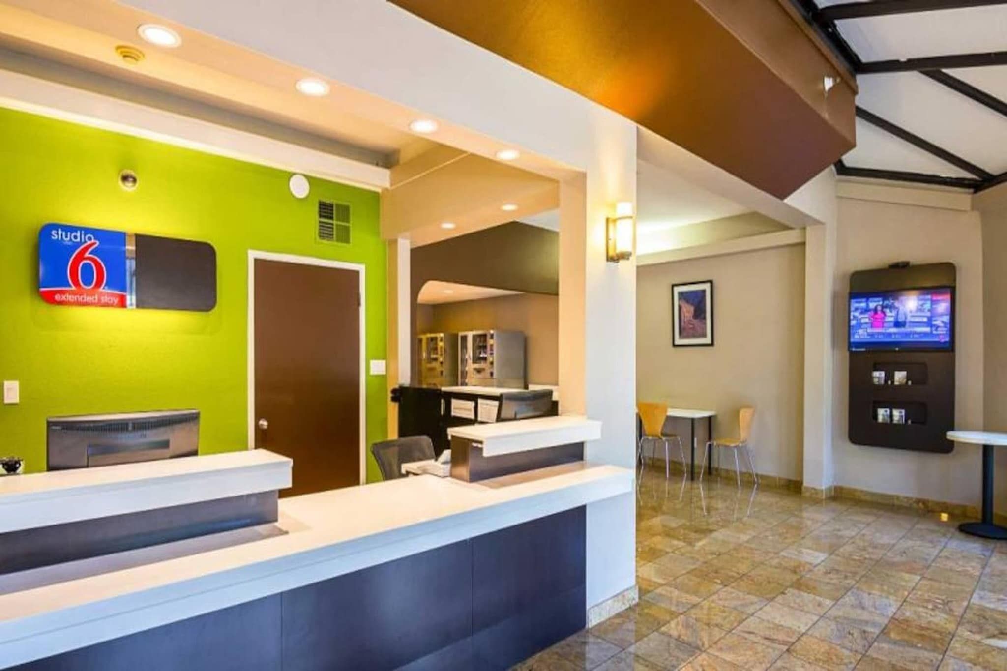 A hotel reception area with a green wall, a reception desk, a television, and a seating area with chairs.