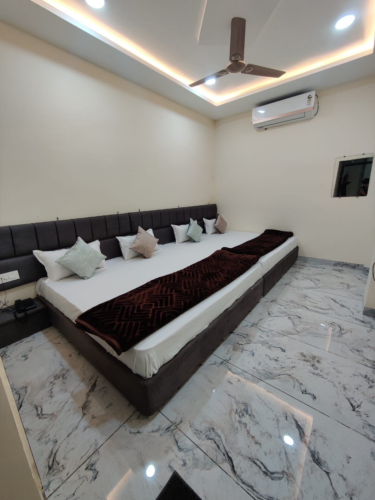 Quarto com cama, mesa de cabeceira, ventilador de teto, aparelho de ar condicionado e um pequeno espelho.
