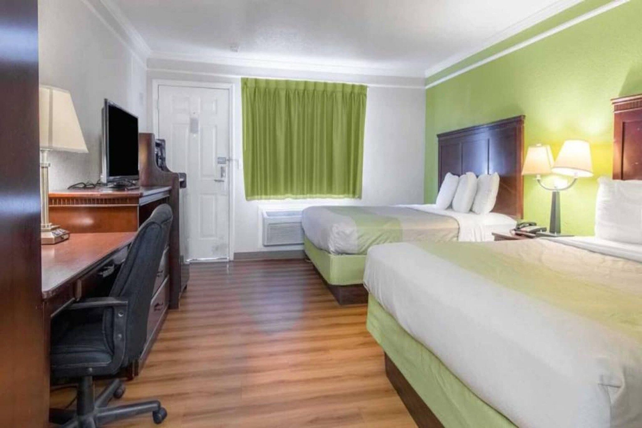 Una habitación de hotel con escritorio, dos camas, una ventana con cortinas verdes y una lámpara de noche.