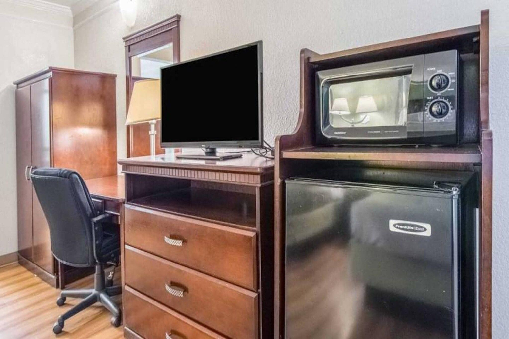 Una habitación compacta con un escritorio de madera, un monitor de computadora, una silla de oficina negra, un refrigerador pequeño y un microondas.