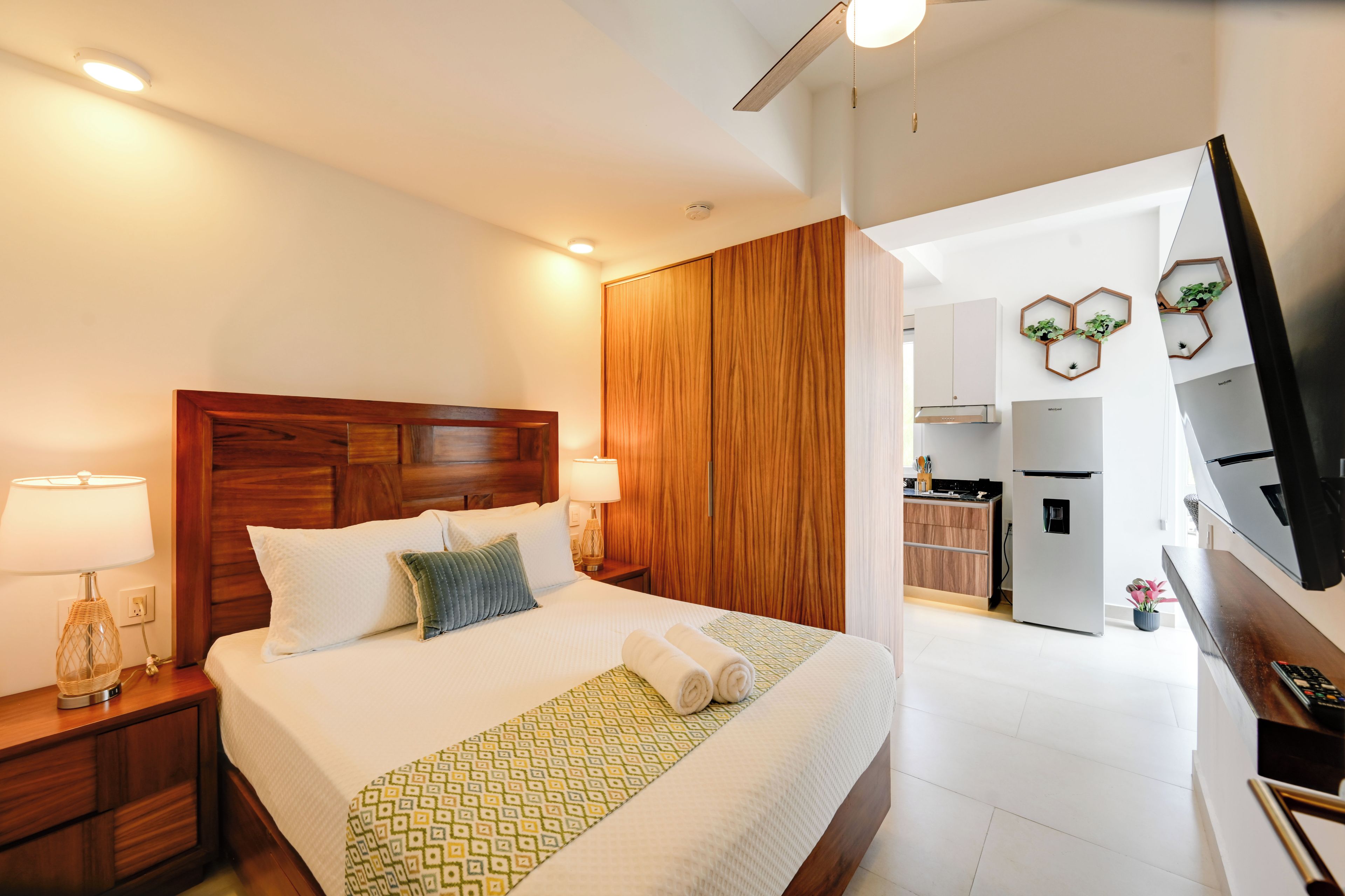 A hotel room with a wooden headboard, a bed with white bedding, a bedside table with a lamp, and a view into the kitchen area.