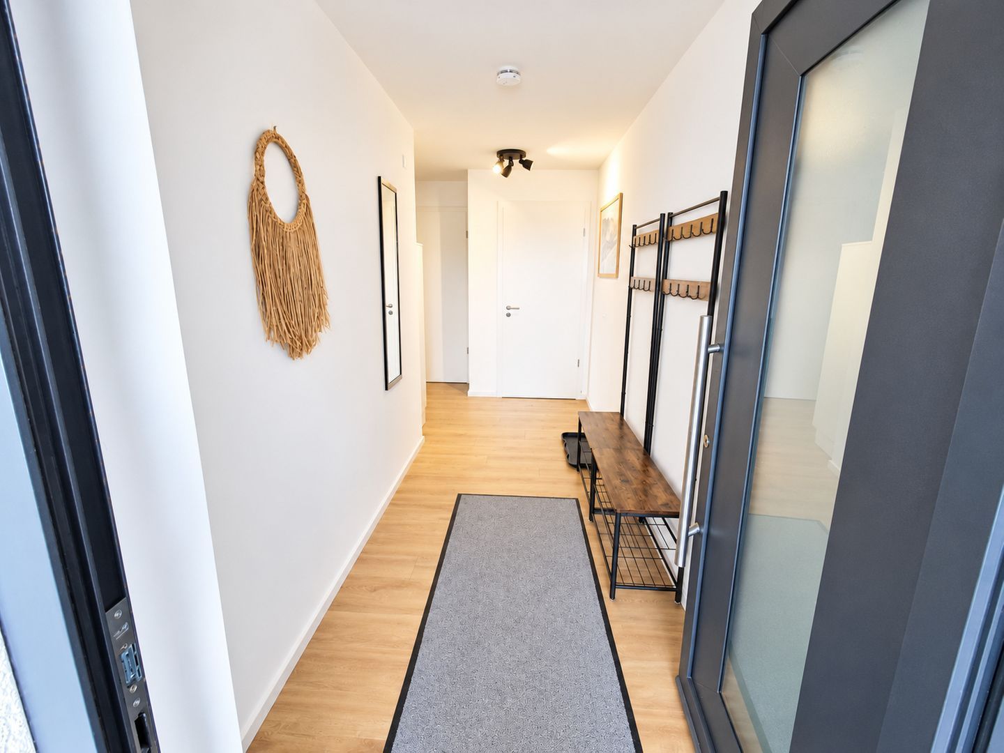 Un couloir doté d’un plancher en bois, d’un banc, d’une décoration murale en macramé et d’une porte au fond.