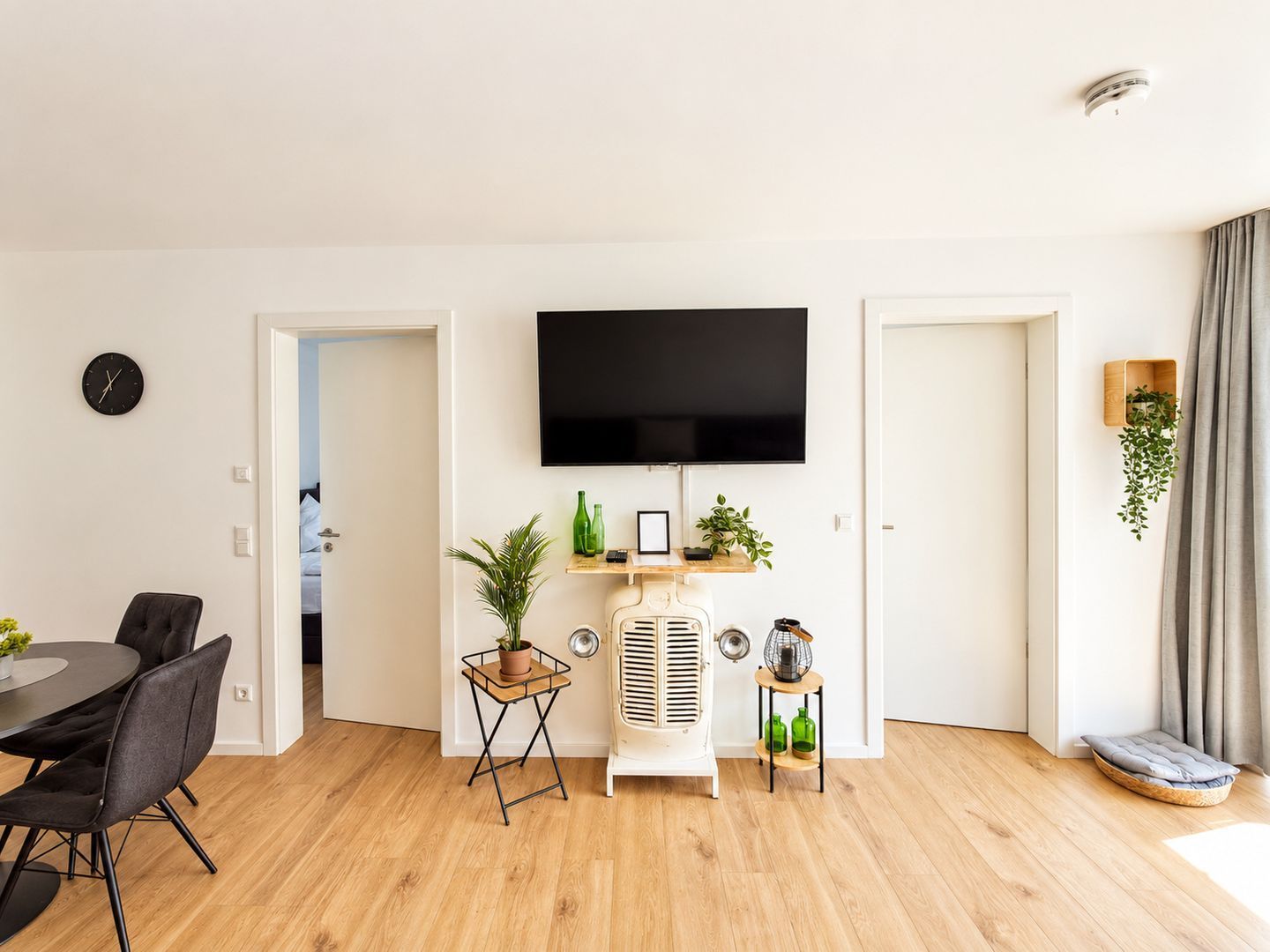 Una sala moderna con televisor de pantalla plana, un comedor con mesa y sillas, un aire acondicionado antiguo y una pequeña planta en un estante.