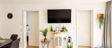 Una sala moderna con televisor de pantalla plana, un comedor con mesa y sillas, un aire acondicionado antiguo y una pequeña planta en un estante.