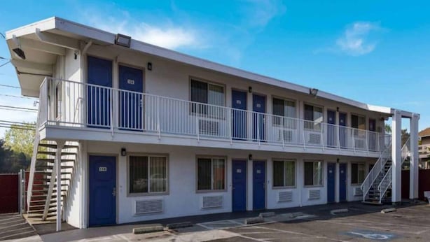 Um motel de dois andares com varanda no andar superior, várias portas e estacionamento em frente.