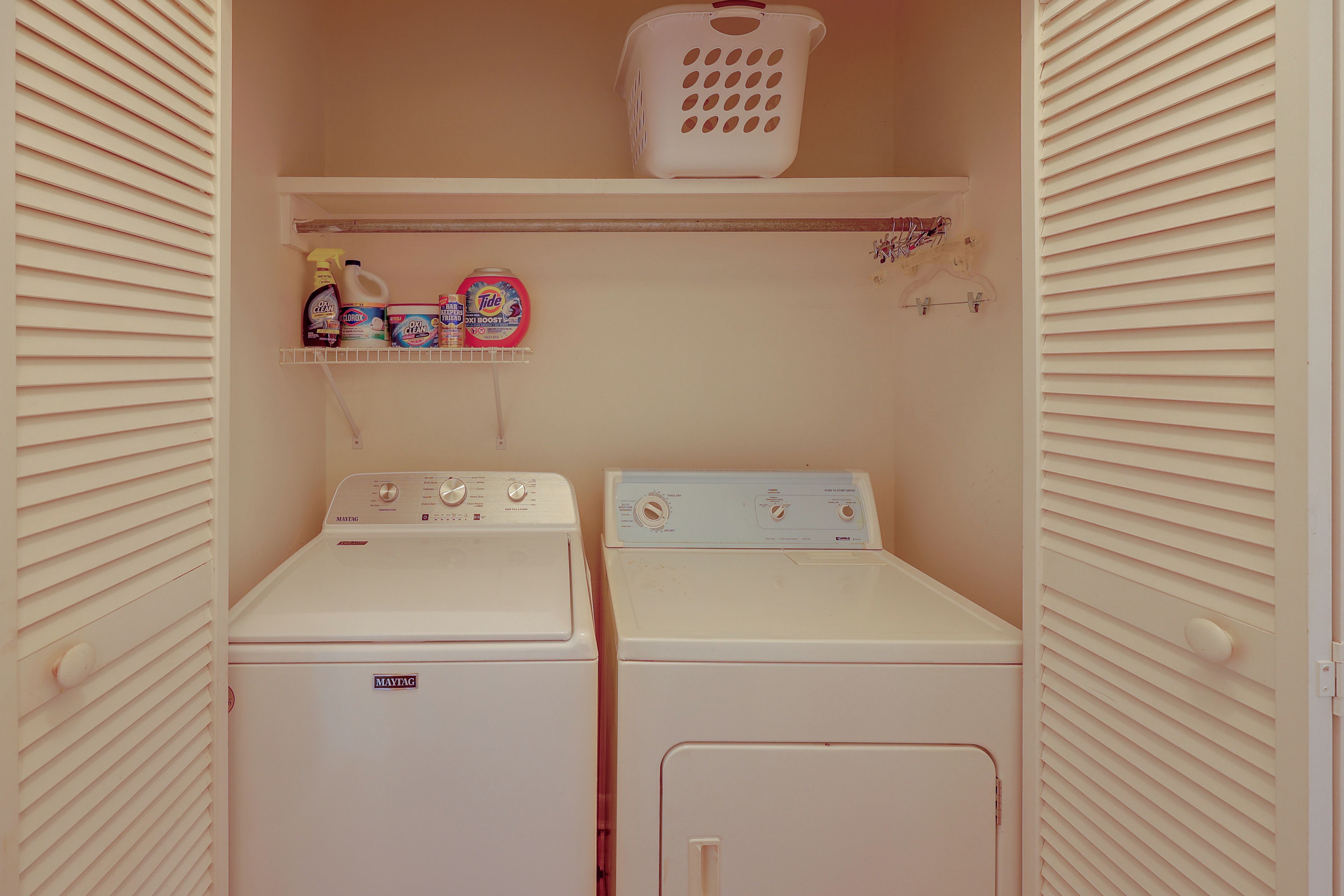 Un cuarto de lavandería con dos lavadoras y secadoras Maytag, un estante con productos de limpieza y una cesta de ropa.