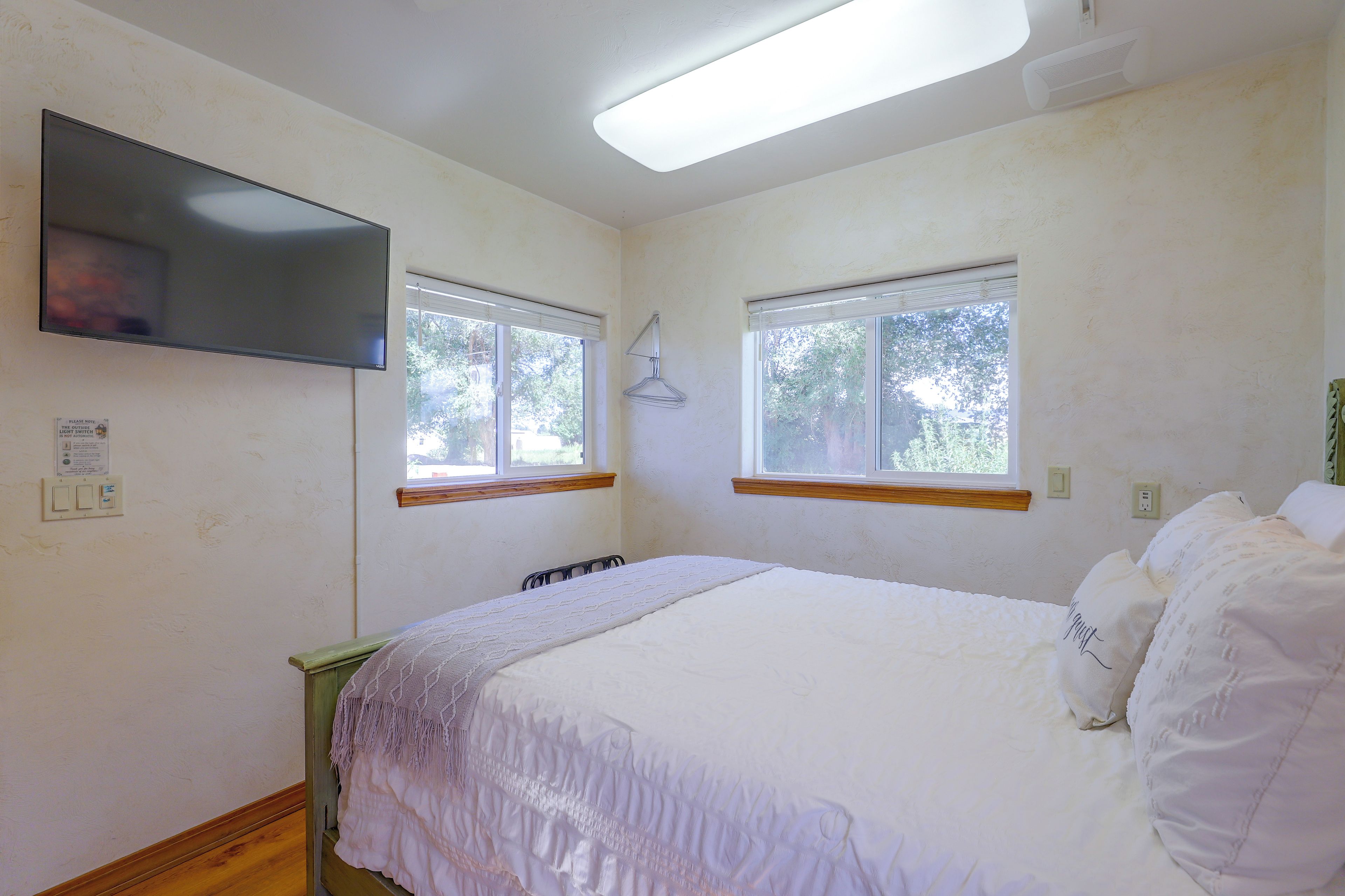A bedroom with a bed, two windows, a flat-screen TV, and a light switch.
