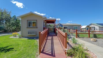 A wooden pathway leading to a single-story house with a covered porch, surrounded by a fenced area and a grassy lawn.