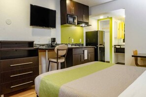 Quarto de hotel com cama, escrivaninha, cadeira, cozinha compacta e televisão.