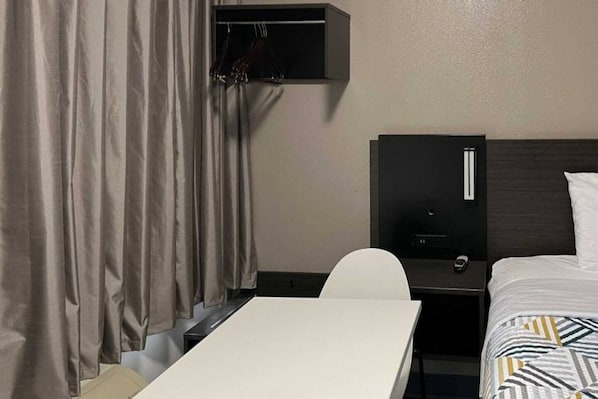 A hotel room with a bed, a desk, and a chair. There is a wardrobe with hanging items and a small shelf.
