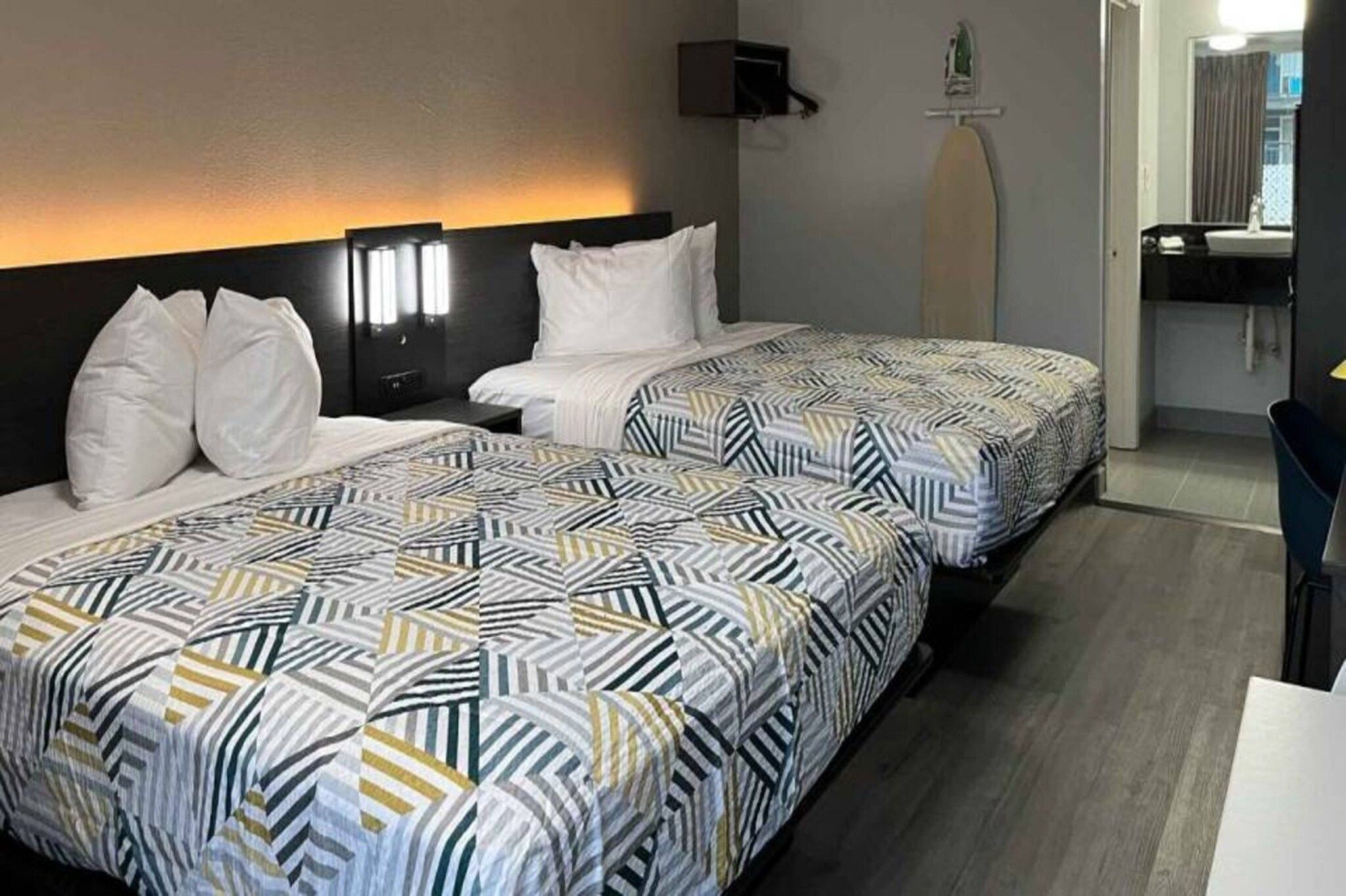 A hotel room with two beds, each with patterned bedding, a headboard, a bedside table, and a wall-mounted lamp.