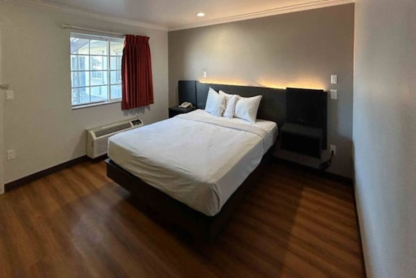 A hotel room with a large bed, a bedside table, a heater, a window with curtains, and a wall-mounted light.